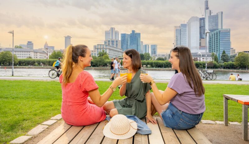 Drie mensen proosten met sinaasappelsap in Frankfurt Sachsenhausen. Op de achtergrond zie je een rivier en de skyline van de stad.