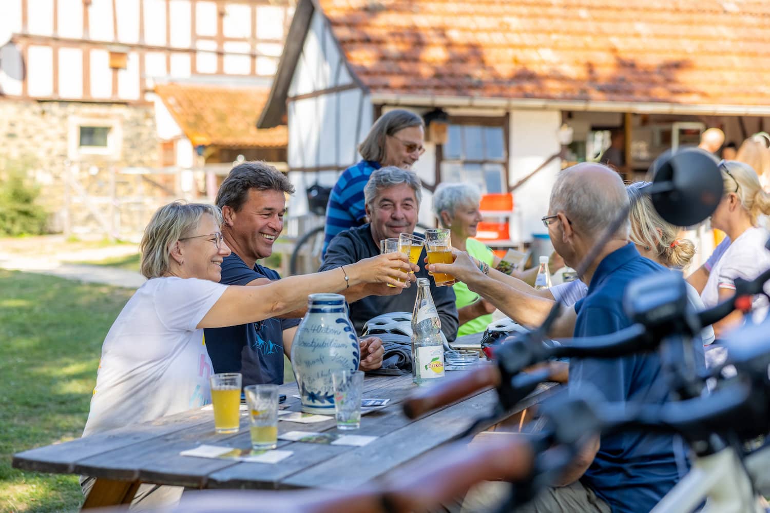 Verschillende mensen zitten buiten aan een tafel voor een vakwerkhuis in de Auenlandhof in Wetterau en proosten met een glas citroensap.