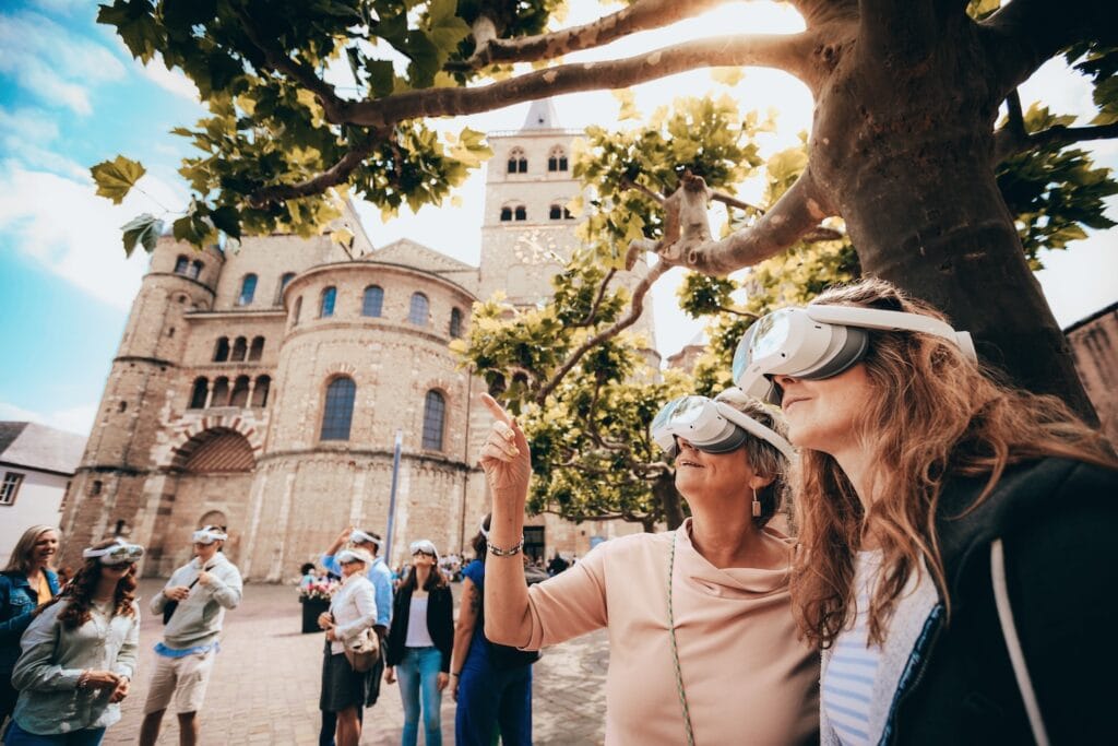 VR-tour 2023: Twee mensen met een VR-bril staan voor de Dom van Trier. Eén persoon wijst omhoog. Andere mensen met een VR-bril staan op de achtergrond