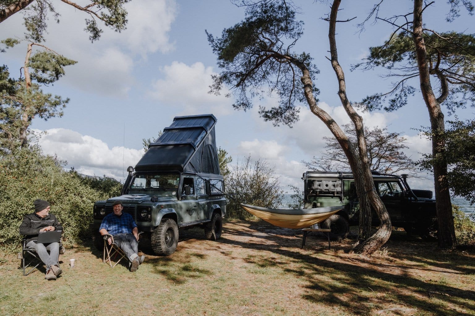 Wildwood Camping op Rügen: twee kampeerders ontspannen naast robuuste offroad-campers onder dennenbomen – puur Vanlife met uitzicht op zee in Duitsland.