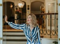 Stijlvolle vrouw met rode lippenstift en elegante outfit opent met een glimlach de glazen deur van een luxueus hotelinterieur. De klassieke kroonluchter, marmeren details en warme sfeer benadrukken de exclusieve charme van het beste luxehotel van Duitsland, waar gastvrijheid en verfijning samenkomen.
