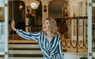 Stijlvolle vrouw met rode lippenstift en elegante outfit opent met een glimlach de glazen deur van een luxueus hotelinterieur. De klassieke kroonluchter, marmeren details en warme sfeer benadrukken de exclusieve charme van het beste luxehotel van Duitsland, waar gastvrijheid en verfijning samenkomen.