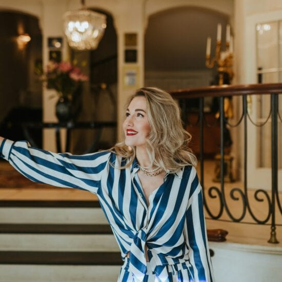 Stijlvolle vrouw met rode lippenstift en elegante outfit opent met een glimlach de glazen deur van een luxueus hotelinterieur. De klassieke kroonluchter, marmeren details en warme sfeer benadrukken de exclusieve charme van het beste luxehotel van Duitsland, waar gastvrijheid en verfijning samenkomen.
