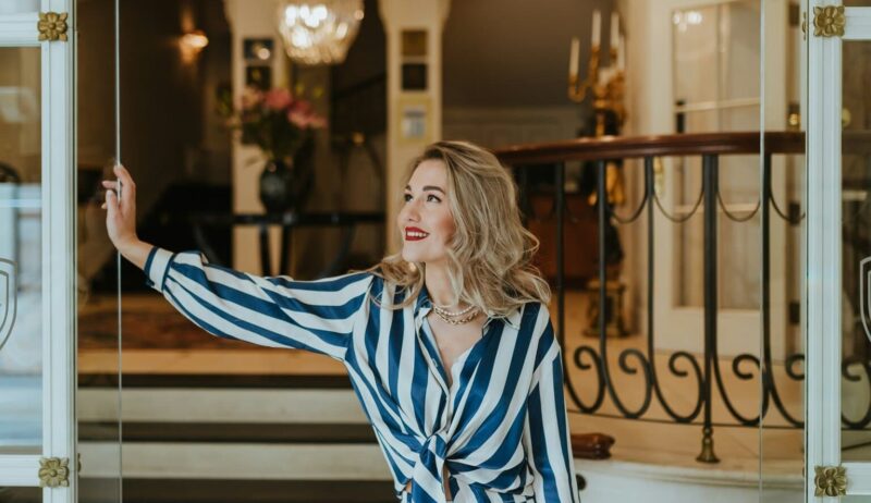 Stijlvolle vrouw met rode lippenstift en elegante outfit opent met een glimlach de glazen deur van een luxueus hotelinterieur. De klassieke kroonluchter, marmeren details en warme sfeer benadrukken de exclusieve charme van het beste luxehotel van Duitsland, waar gastvrijheid en verfijning samenkomen.