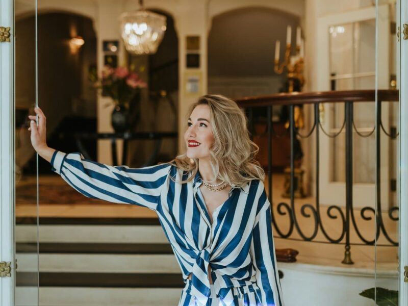 Stijlvolle vrouw met rode lippenstift en elegante outfit opent met een glimlach de glazen deur van een luxueus hotelinterieur. De klassieke kroonluchter, marmeren details en warme sfeer benadrukken de exclusieve charme van het beste luxehotel van Duitsland, waar gastvrijheid en verfijning samenkomen.