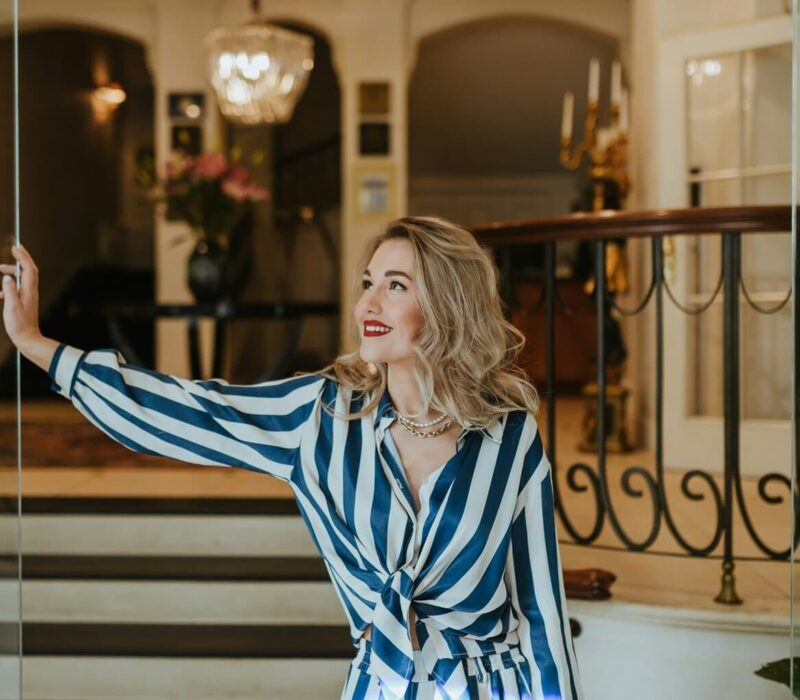 Stijlvolle vrouw met rode lippenstift en elegante outfit opent met een glimlach de glazen deur van een luxueus hotelinterieur. De klassieke kroonluchter, marmeren details en warme sfeer benadrukken de exclusieve charme van het beste luxehotel van Duitsland, waar gastvrijheid en verfijning samenkomen.