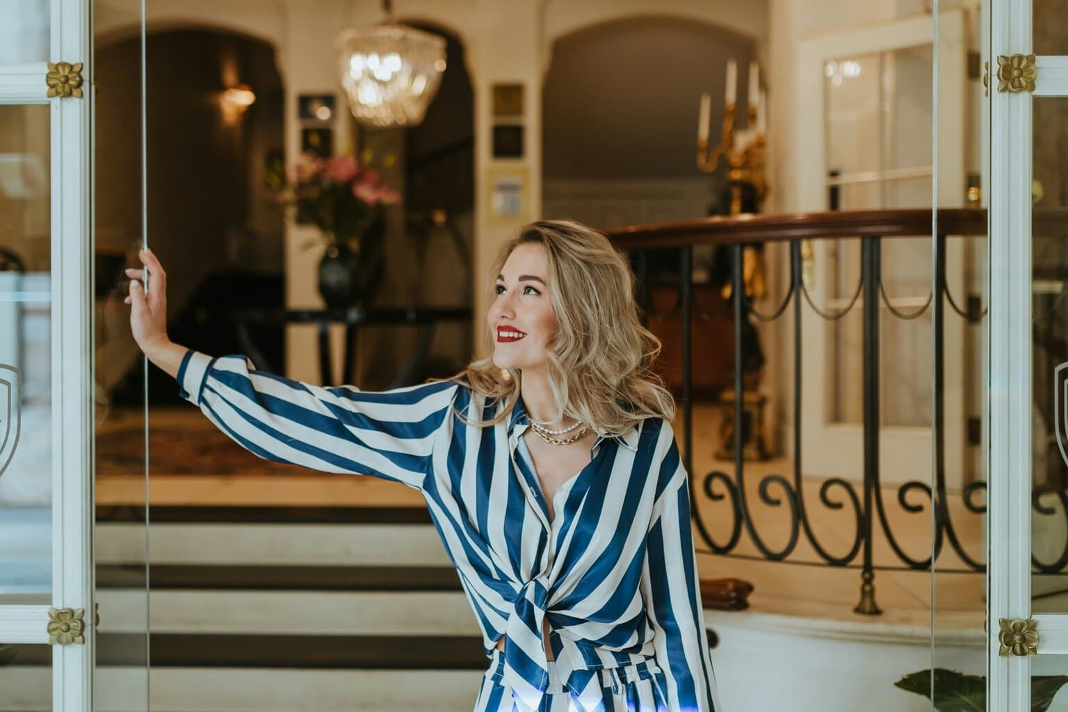 Stijlvolle vrouw met rode lippenstift en elegante outfit opent met een glimlach de glazen deur van een luxueus hotelinterieur. De klassieke kroonluchter, marmeren details en warme sfeer benadrukken de exclusieve charme van het beste luxehotel van Duitsland, waar gastvrijheid en verfijning samenkomen.