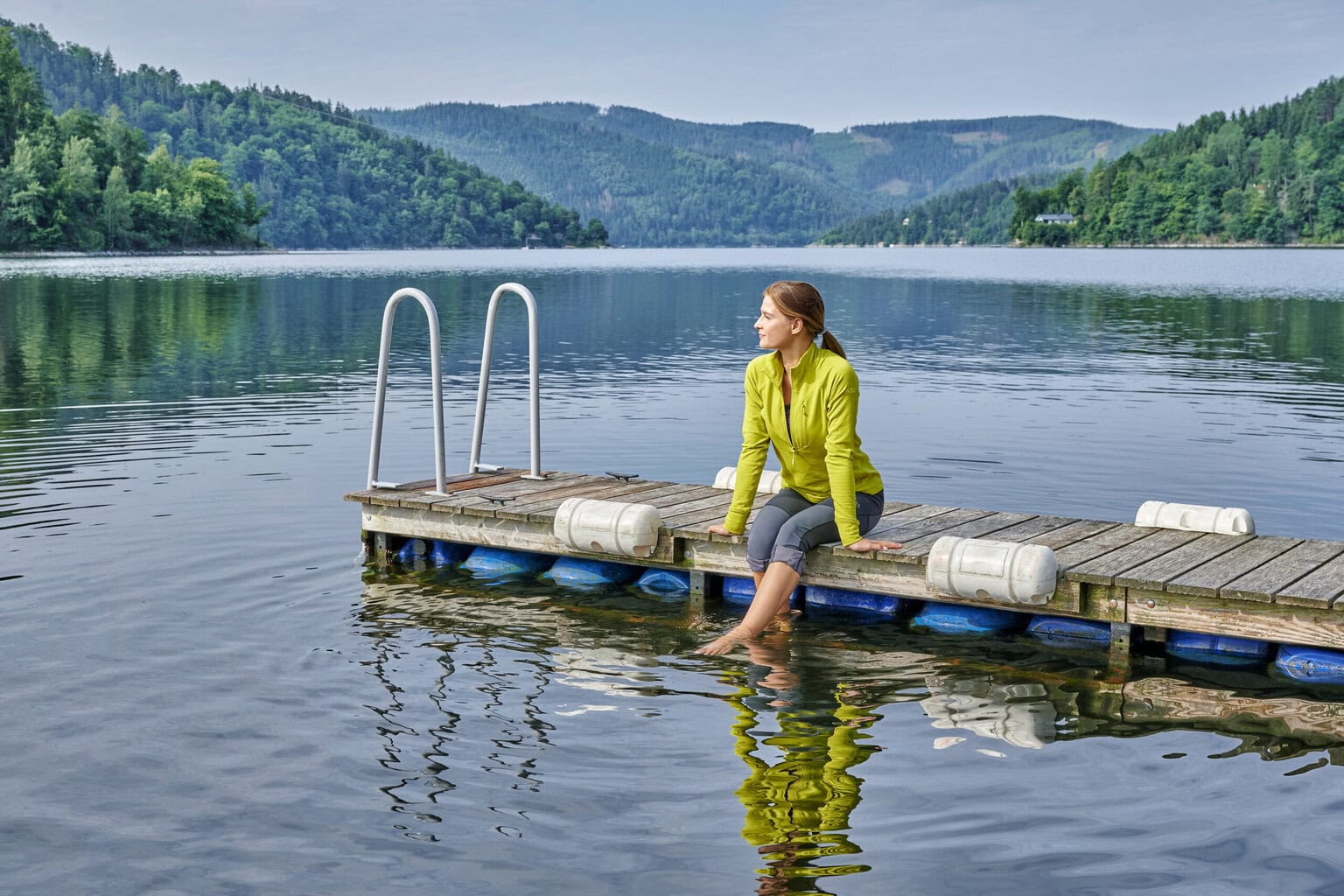 Vrouw geniet van de rust aan het Thüringer Meer, zittend op een houten steiger in het Thüringer Woud, omgeven door uitgestrekte bossen, berglandschap en spiegelend water – een perfecte plek voor natuurliefhebbers.