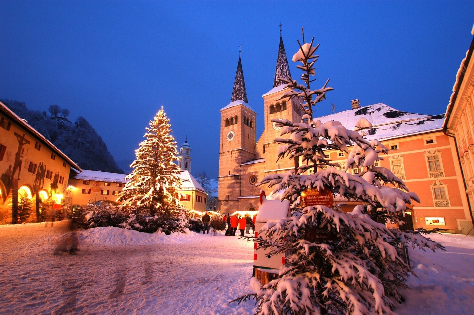 Verlichte kerstboom en versierde kraampjes op de sfeervolle kerstmarkt van Berchtesgaden in Beieren, met besneeuwde daken en de torens van de Stiftskerk op de achtergrond. Een typisch winters tafereel in de Alpen met kerstversiering en historische gebouwen.