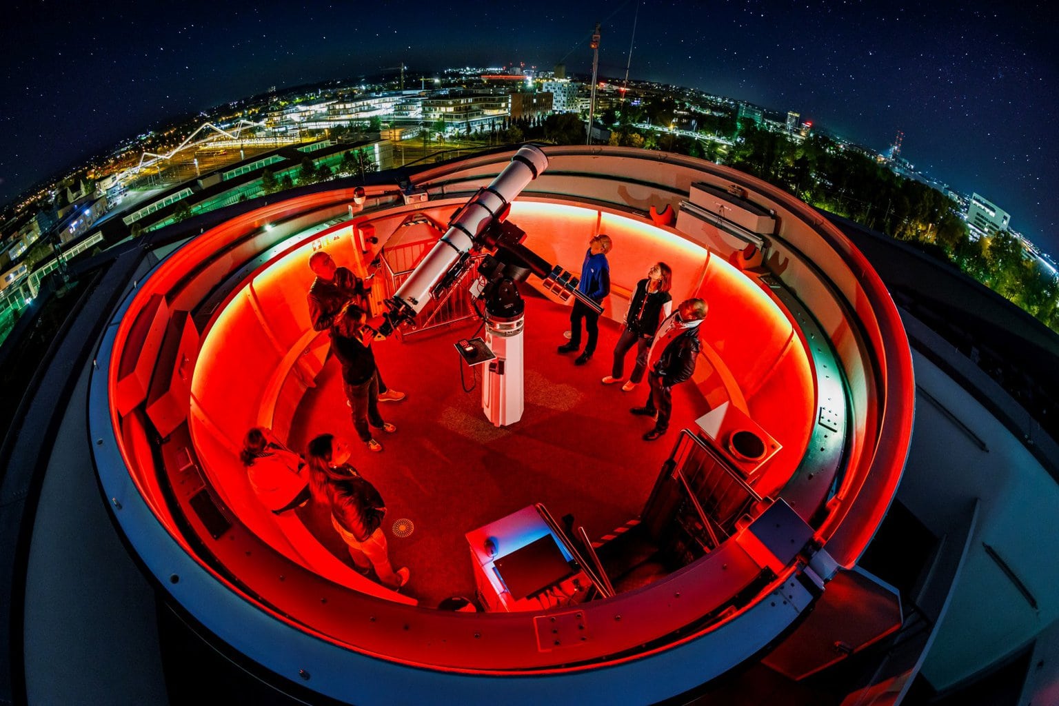 Groep bezoekers rond telescoop in rood verlichte sterrenwacht tijdens zuiden night, met uitzicht op de stad en een heldere sterrenhemel vol fonkelende lichten.