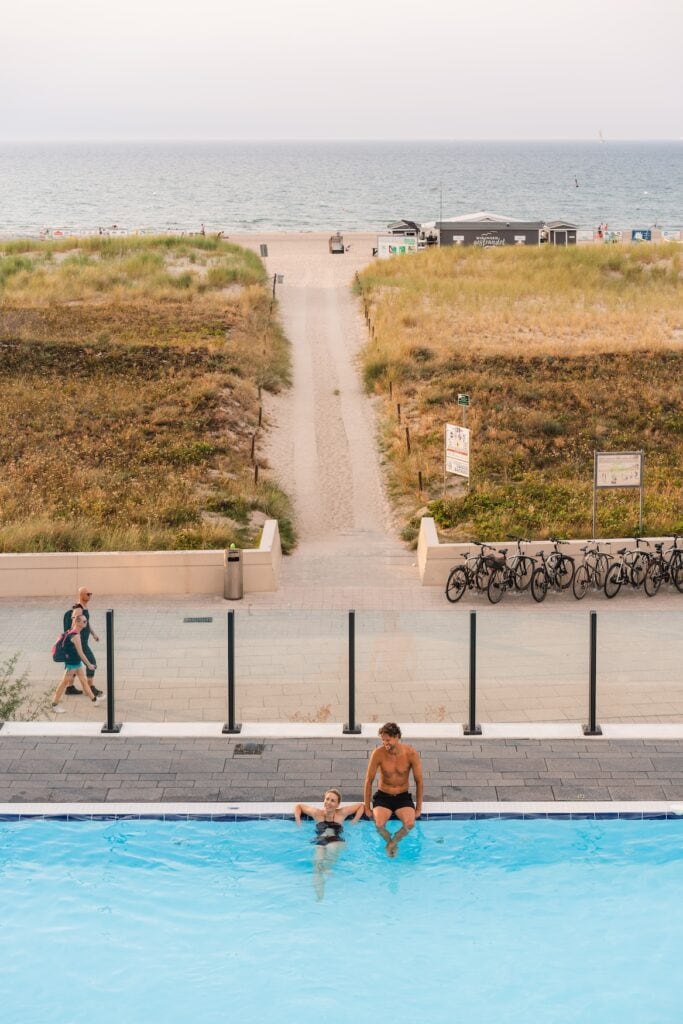 Twee personen ontspannen zich in het zwembad, dat zich buiten bij het Hotel aja Warnemünde bevindt. Op de achtergrond zie je het strand van de Oostzee.