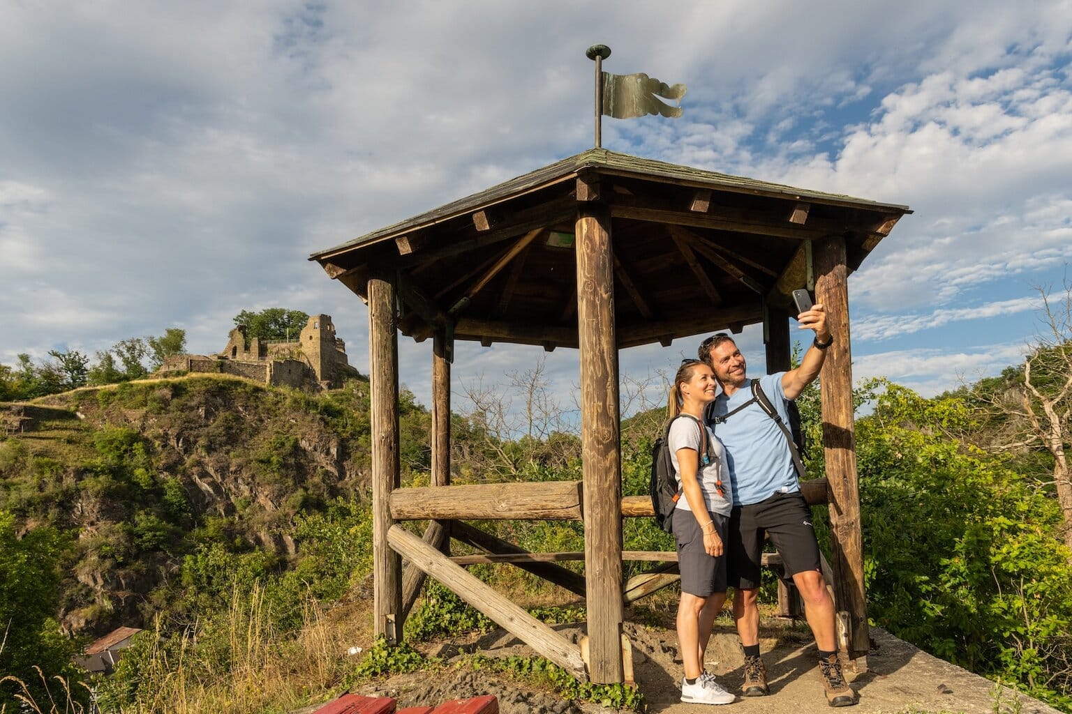 Een wandelend paar maakt een selfie bij een uitkijkpunt langs de AhrSteig in het Ahrdal, met uitzicht op een middeleeuwse burchtruïne. De route combineert natuur, panorama’s en cultuur en behoort tot de mooiste wandelpaden in het Ahrdal.