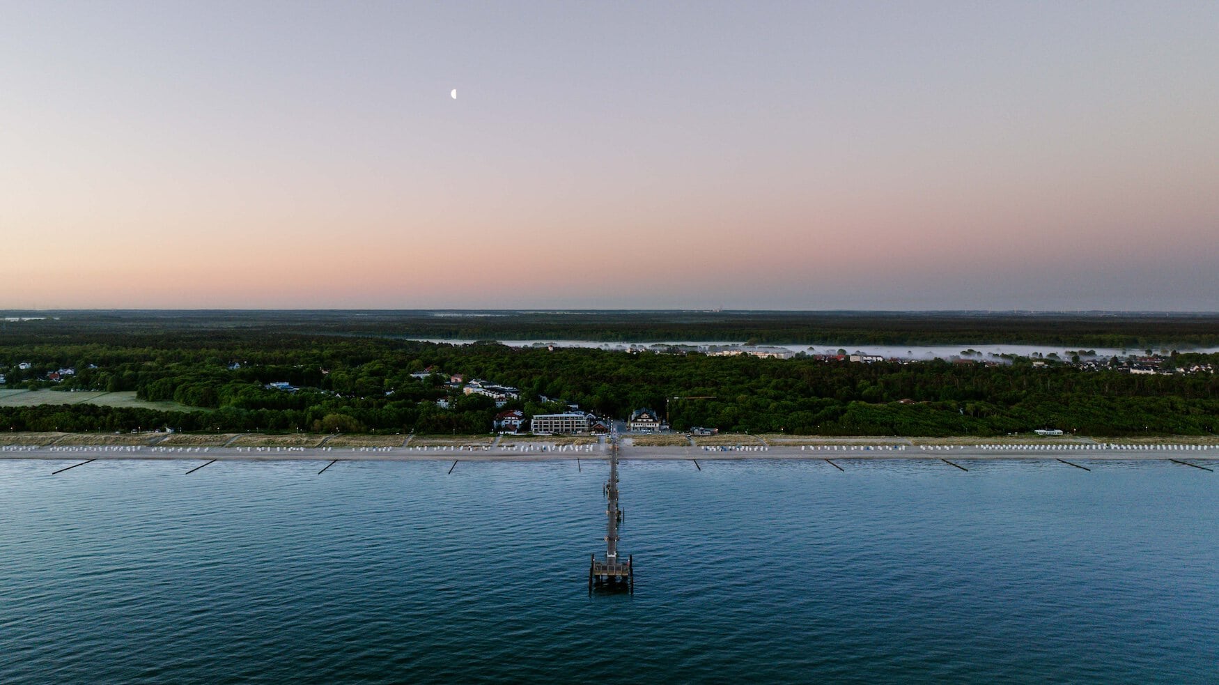 Avondstemming boven de Oostzee bij Graal-Müritz: de Seebrücke steekt in zee uit terwijl de maan boven het kustbos staat.