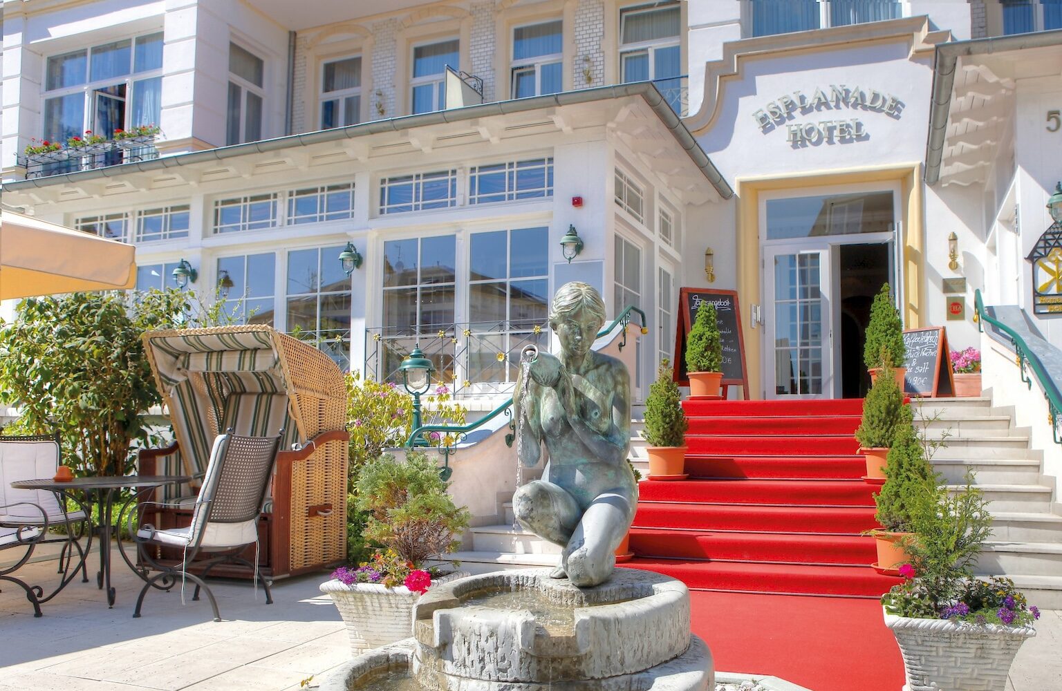 Voor de ingang van het Esplanade Hotel staat een fontein met een standbeeld van een vrouw die water uitgiet. Een trap, versierd met een rood tapijt en planten, leidt naar de witte gevel met glazen deur.