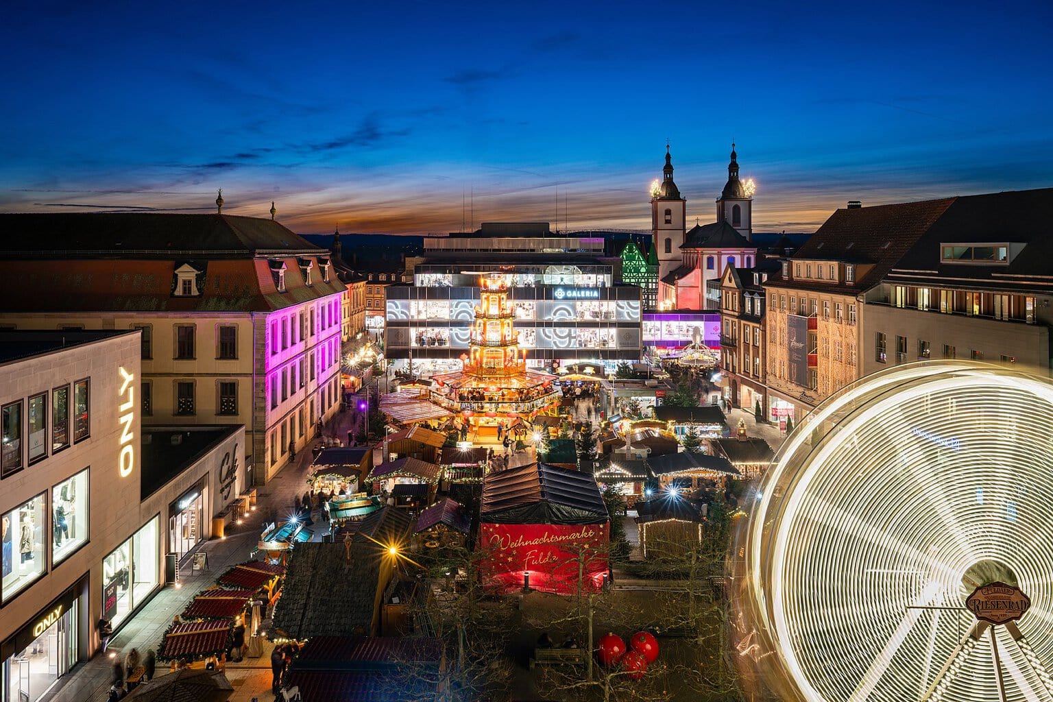 De kerstmarkt in Fulda van bovenaf met kerstpiramide, verlicht reuzenrad en verlichte kerktorens bij schemering