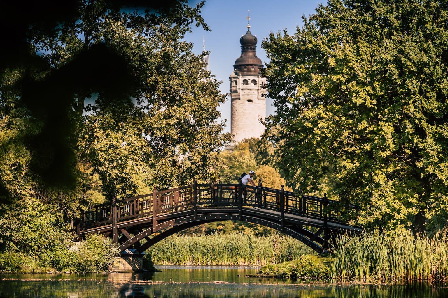 Een fietser rijdt over de brug in het Johannapark in Leipzig. Op de achtergrond is een toren van het Nieuwe Stadhuis te zien. Groene bomen steken uit boven de oever van de rivier de Pleiße