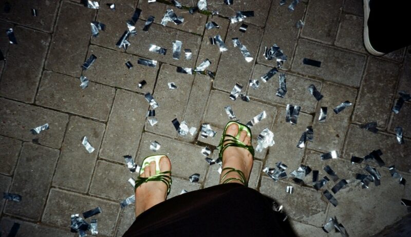 Glinsterend confetti op straat na een Stuttgart party, waar dansende voeten in groene sandalen herinneren aan de nacht vol ritme en vrijheid.