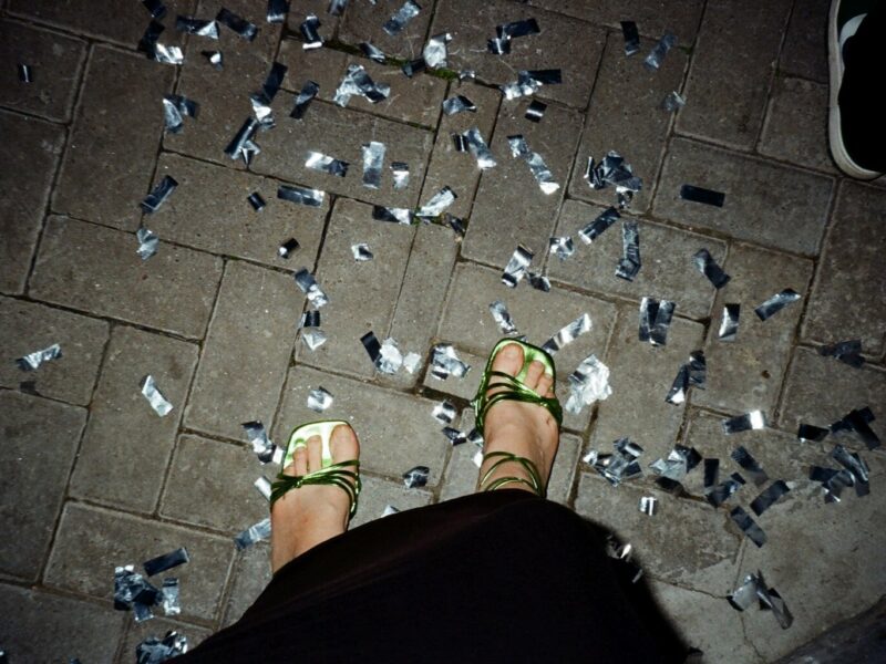 Glinsterend confetti op straat na een Stuttgart party, waar dansende voeten in groene sandalen herinneren aan de nacht vol ritme en vrijheid.