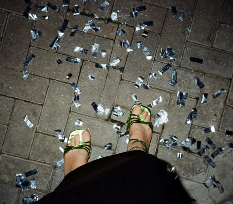 Glinsterend confetti op straat na een Stuttgart party, waar dansende voeten in groene sandalen herinneren aan de nacht vol ritme en vrijheid.