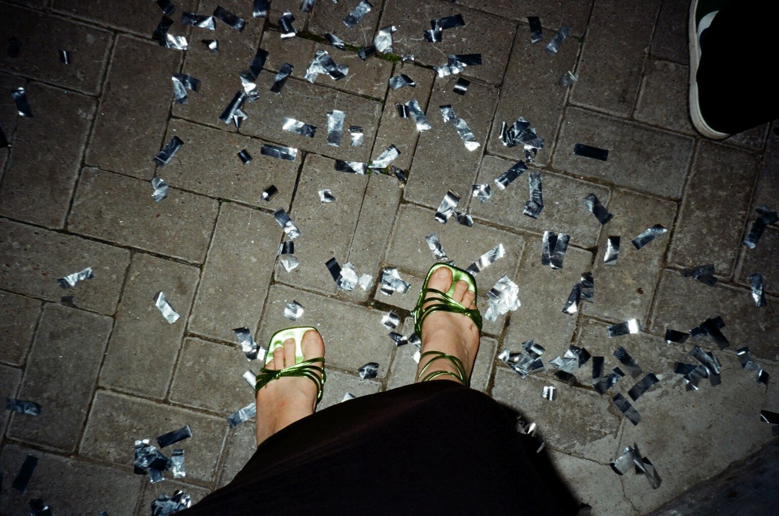 Glinsterend confetti op straat na een Stuttgart party, waar dansende voeten in groene sandalen herinneren aan de nacht vol ritme en vrijheid.