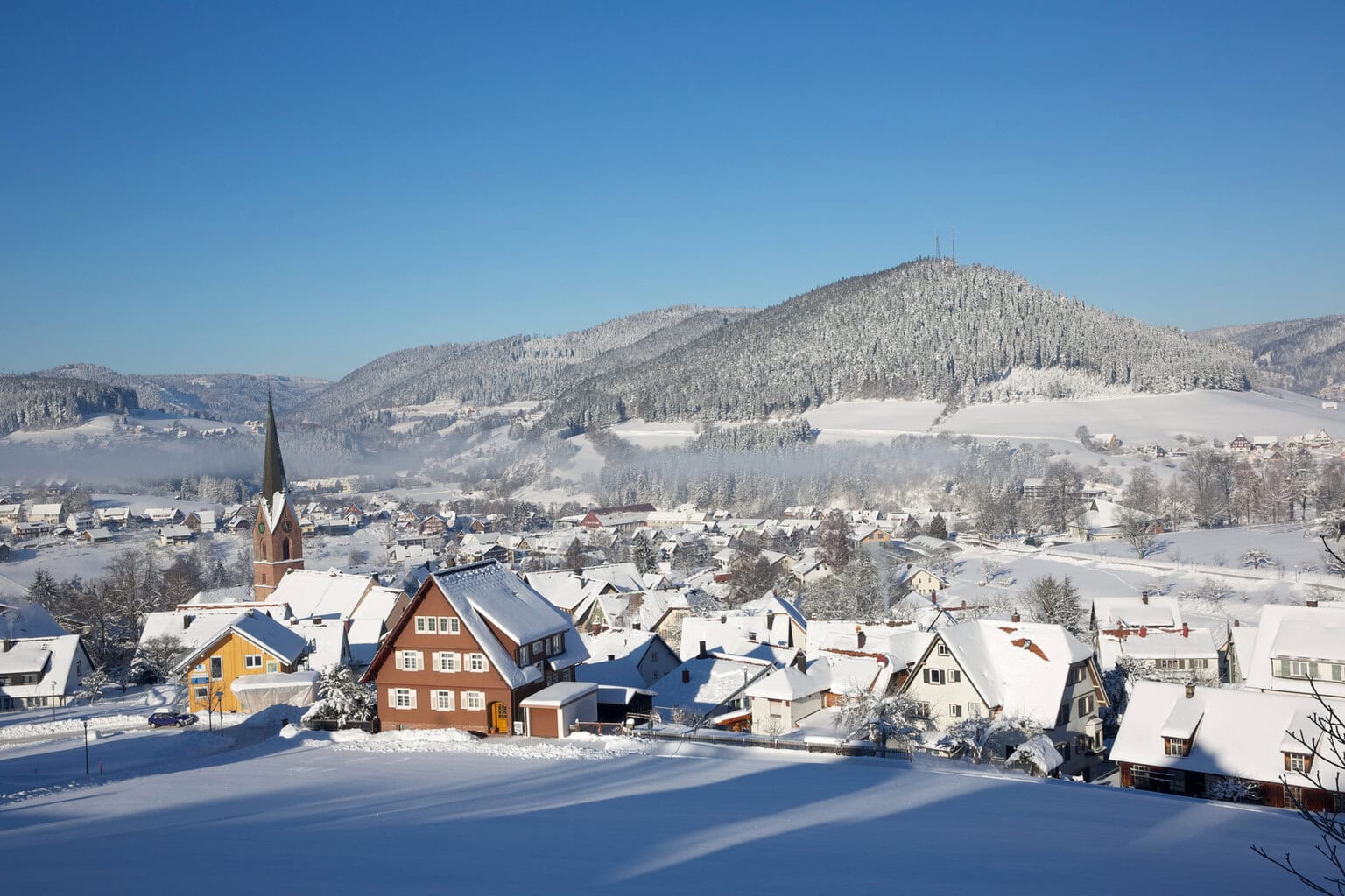 Onder een strakblauwe hemel glinsteren de daken van een bergdorp in de winterzon – pure rust en betoverend Sneeuwplezier Zwarte Woud.
