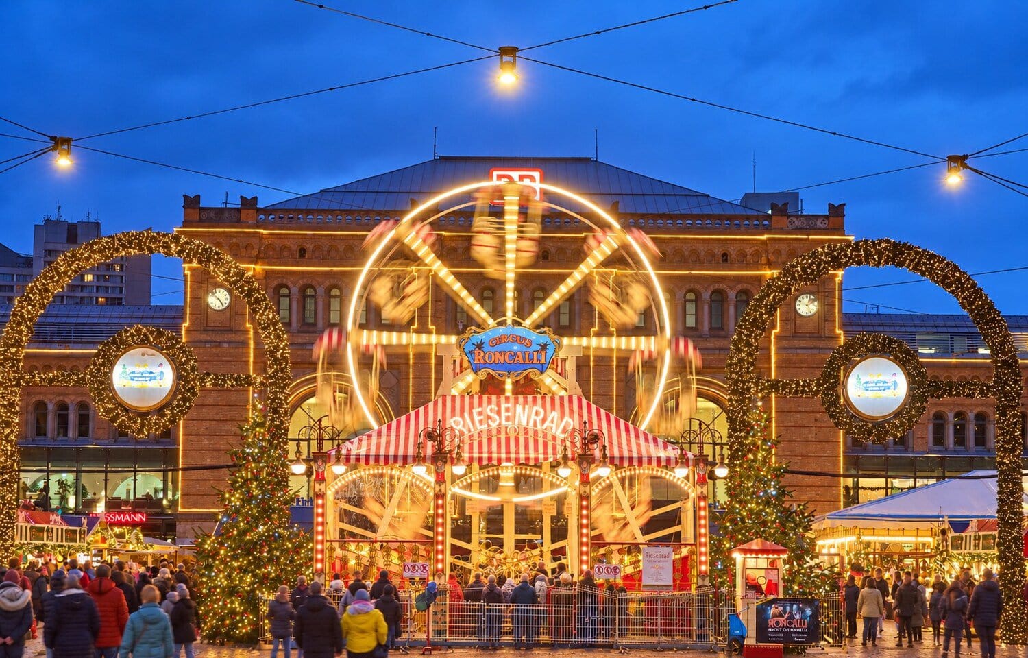 Verlicht reuzenrad van het Roncalli-kerstdorp voor het Hauptbahnhof van Hannover tijdens het blauwe uur, met lichtbogen, kerstbomen en bezoekers op de Ernst-August-Platz
