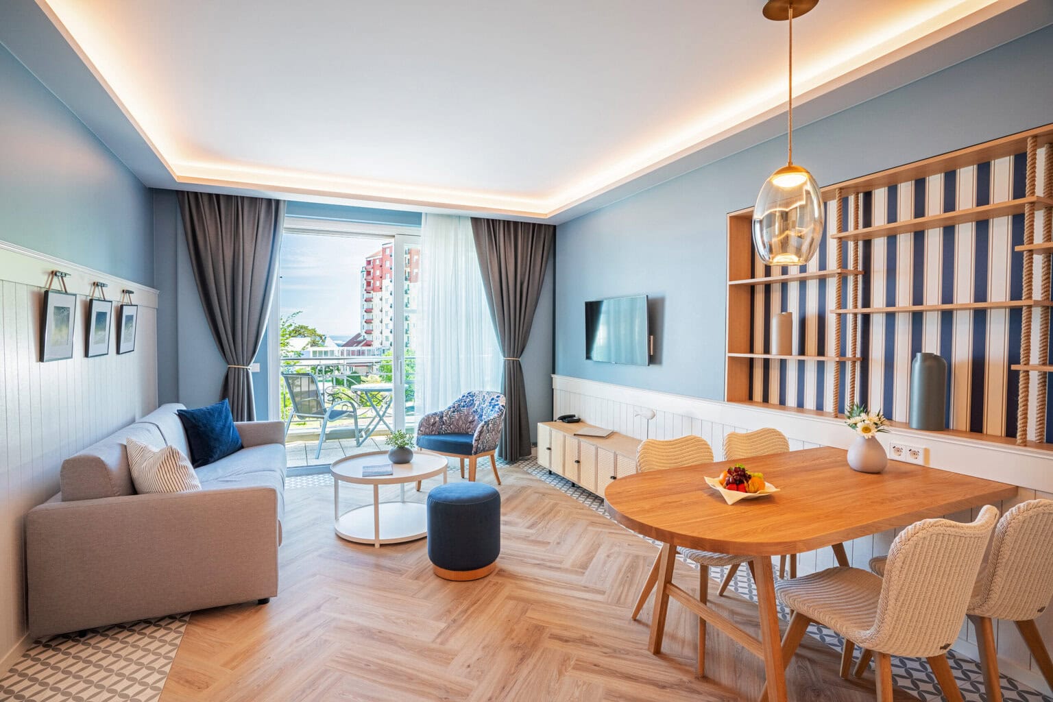 Moderne hotelkamer in de Ostseeresidenz Heringsdorf met houten tafel en stoelen, wandplank, tv-kast, televisie, bank en fauteuil. Op de achtergrond het balkon en uitzicht op Heringsdorf