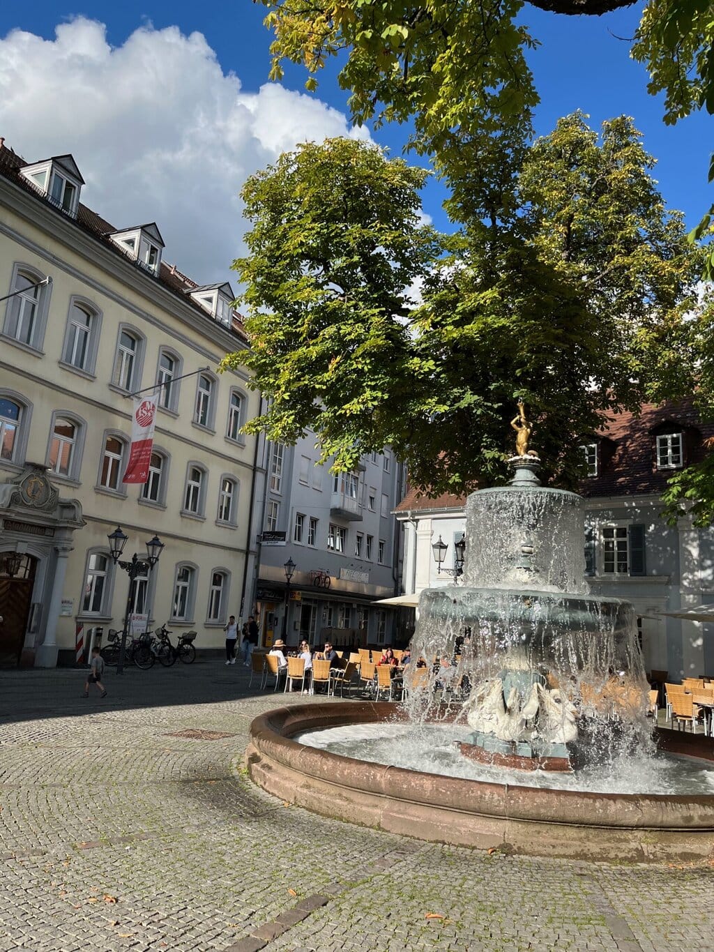 Zonnestralen vallen op een levendig plein met fontein en terrasjes, waar sfeer en historisch charme samen de smaken van Saarland Rijnland-Palts omringen.