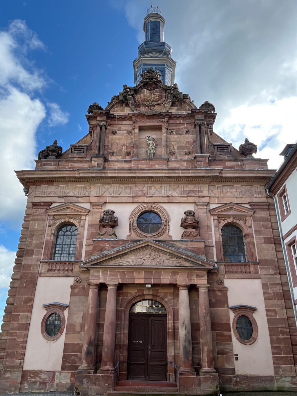 Een barokke kerkgevel in warm rood zandsteen rijst statig op onder een blauwe lucht, een architectonisch juweel midden in Saarland Rijnland-Palts.