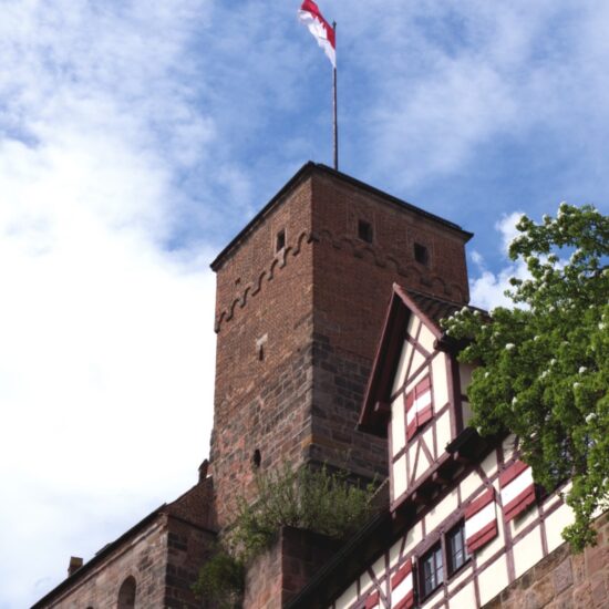Uitzicht op de historische toren van de Kaiserburg in Neurenberg, met vakwerkhuizen en een wapperende rood-witte vlag onder een blauwe hemel. Een sfeervol hoogtepunt voor activiteiten in Neurenberg tijdens een weekendje weg.