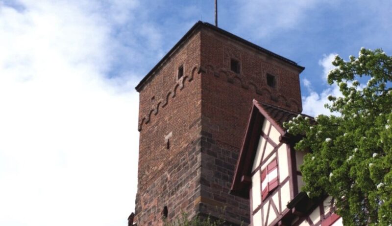 Uitzicht op de historische toren van de Kaiserburg in Neurenberg, met vakwerkhuizen en een wapperende rood-witte vlag onder een blauwe hemel. Een sfeervol hoogtepunt voor activiteiten in Neurenberg tijdens een weekendje weg.