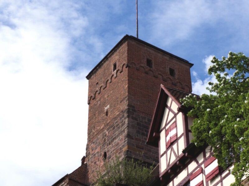 Uitzicht op de historische toren van de Kaiserburg in Neurenberg, met vakwerkhuizen en een wapperende rood-witte vlag onder een blauwe hemel. Een sfeervol hoogtepunt voor activiteiten in Neurenberg tijdens een weekendje weg.