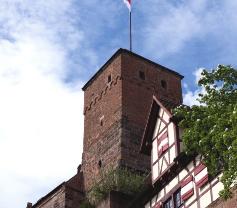 Uitzicht op de historische toren van de Kaiserburg in Neurenberg, met vakwerkhuizen en een wapperende rood-witte vlag onder een blauwe hemel. Een sfeervol hoogtepunt voor activiteiten in Neurenberg tijdens een weekendje weg.
