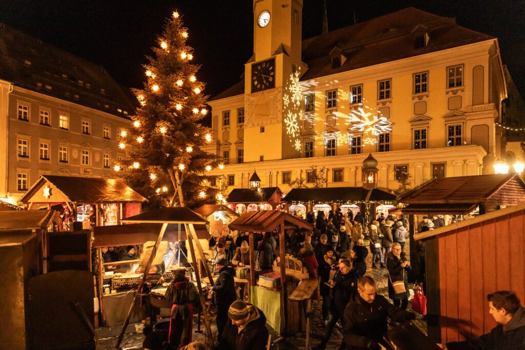 Bezoekers tussen houten kramen op de Wenzelsmarkt in Bautzen, met kerstboom en het stadhuis met sneeuwvlokprojectie