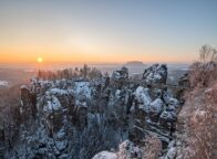 Besneeuwde Basteibrug bij zonsopkomst in de Sächsische Schweiz: winterse rotsformaties en diepe kloven maken Wandelen in Saksisch Zwitserland tot een magische ervaring.