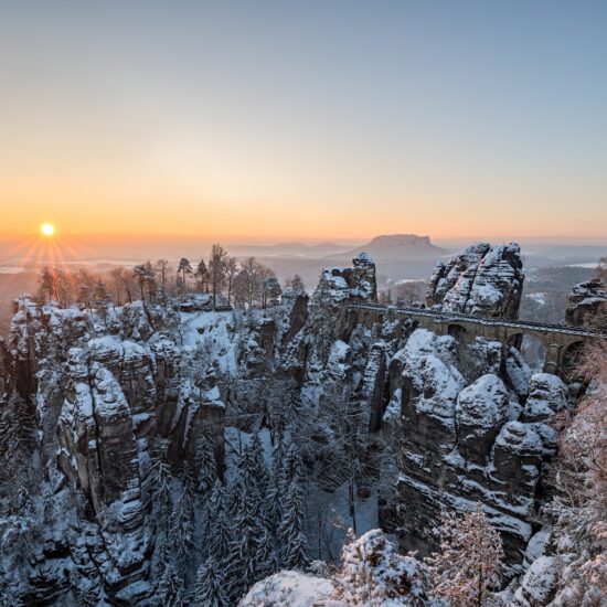 Besneeuwde Basteibrug bij zonsopkomst in de Sächsische Schweiz: winterse rotsformaties en diepe kloven maken Wandelen in Saksisch Zwitserland tot een magische ervaring.