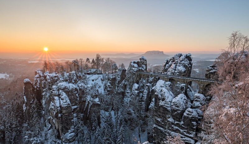 Besneeuwde Basteibrug bij zonsopkomst in de Sächsische Schweiz: winterse rotsformaties en diepe kloven maken Wandelen in Saksisch Zwitserland tot een magische ervaring.