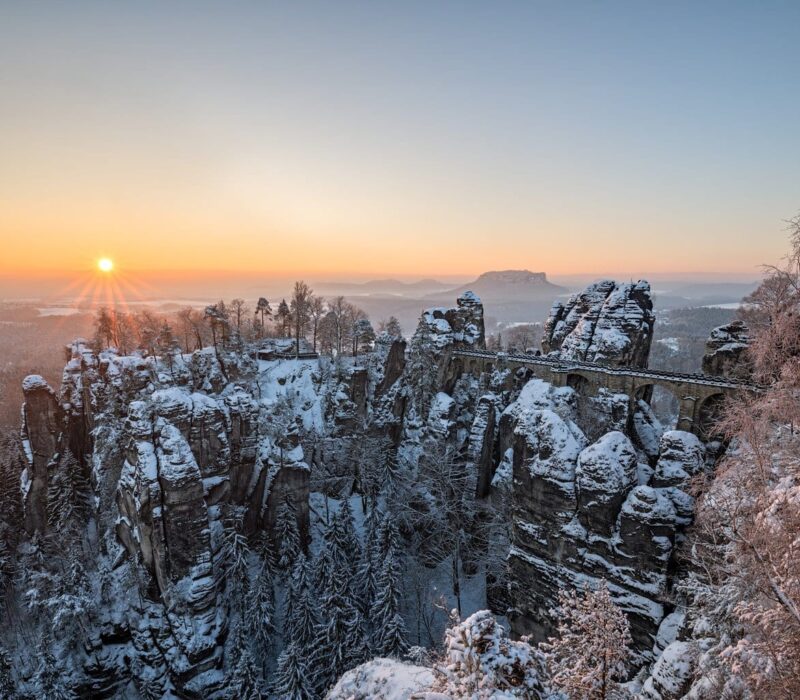 Besneeuwde Basteibrug bij zonsopkomst in de Sächsische Schweiz: winterse rotsformaties en diepe kloven maken Wandelen in Saksisch Zwitserland tot een magische ervaring.