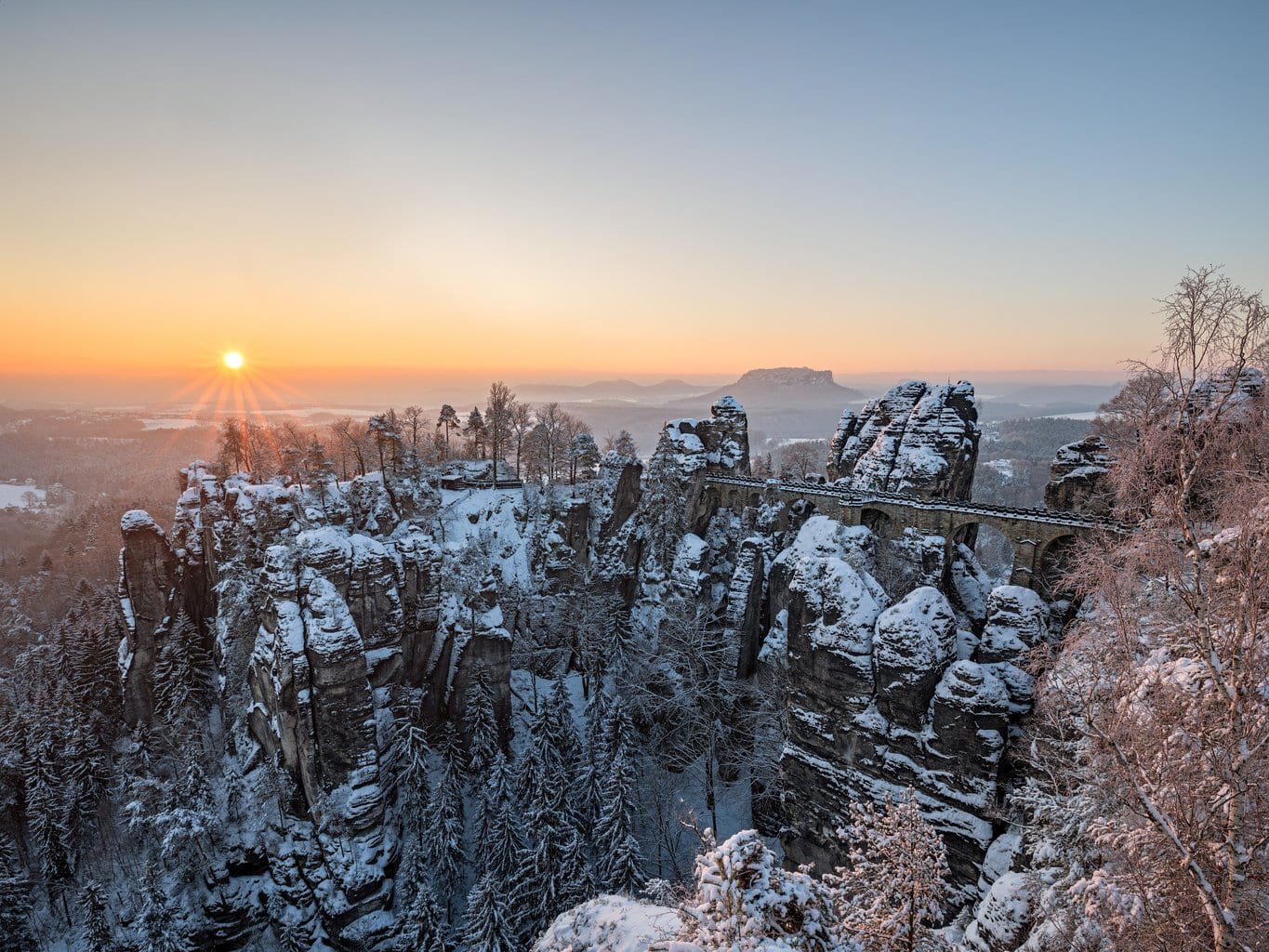 Besneeuwde Basteibrug bij zonsopkomst in de Sächsische Schweiz: winterse rotsformaties en diepe kloven maken Wandelen in Saksisch Zwitserland tot een magische ervaring.
