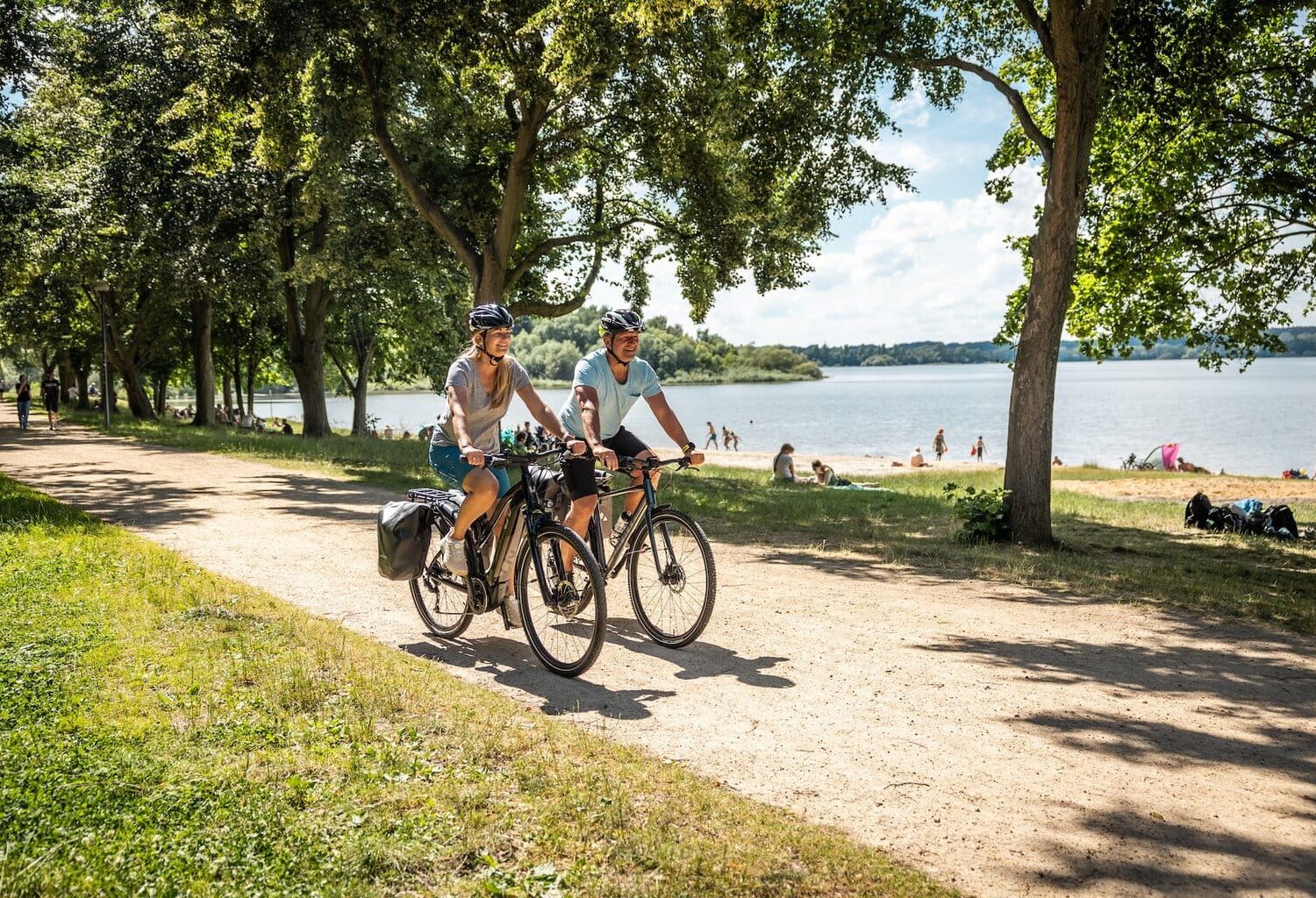 Twee fietsers rijden over het fietspad langs het strand van het stuwmeer in de fietsregio Bautzen. Op de achtergrond zie je het stuwmeer en mensen die op het strand van de zon genieten.