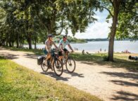Twee fietsers rijden over het fietspad langs het strand van het stuwmeer in de fietsregio Bautzen. Op de achtergrond zie je het stuwmeer en mensen die op het strand van de zon genieten.
