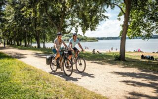 Twee fietsers rijden over het fietspad langs het strand van het stuwmeer in de fietsregio Bautzen. Op de achtergrond zie je het stuwmeer en mensen die op het strand van de zon genieten.