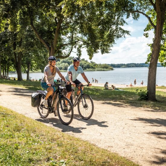 Twee fietsers rijden over het fietspad langs het strand van het stuwmeer in de fietsregio Bautzen. Op de achtergrond zie je het stuwmeer en mensen die op het strand van de zon genieten.