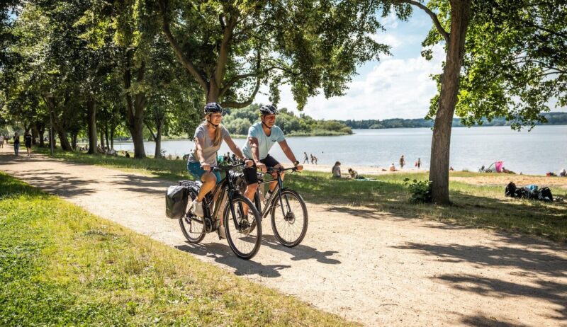 Twee fietsers rijden over het fietspad langs het strand van het stuwmeer in de fietsregio Bautzen. Op de achtergrond zie je het stuwmeer en mensen die op het strand van de zon genieten.