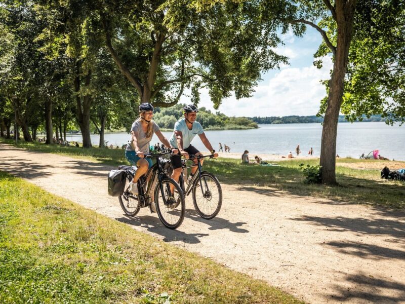 Twee fietsers rijden over het fietspad langs het strand van het stuwmeer in de fietsregio Bautzen. Op de achtergrond zie je het stuwmeer en mensen die op het strand van de zon genieten.
