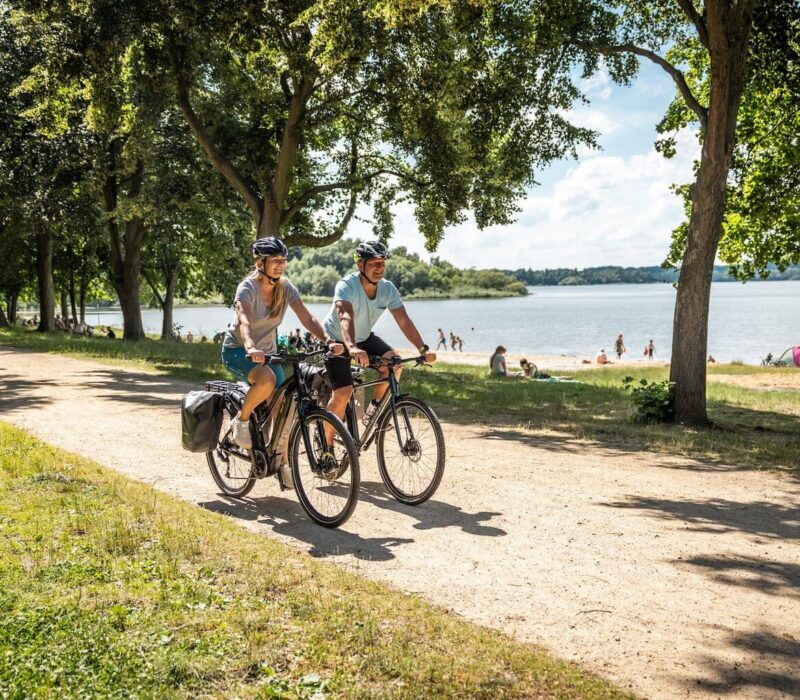 Twee fietsers rijden over het fietspad langs het strand van het stuwmeer in de fietsregio Bautzen. Op de achtergrond zie je het stuwmeer en mensen die op het strand van de zon genieten.