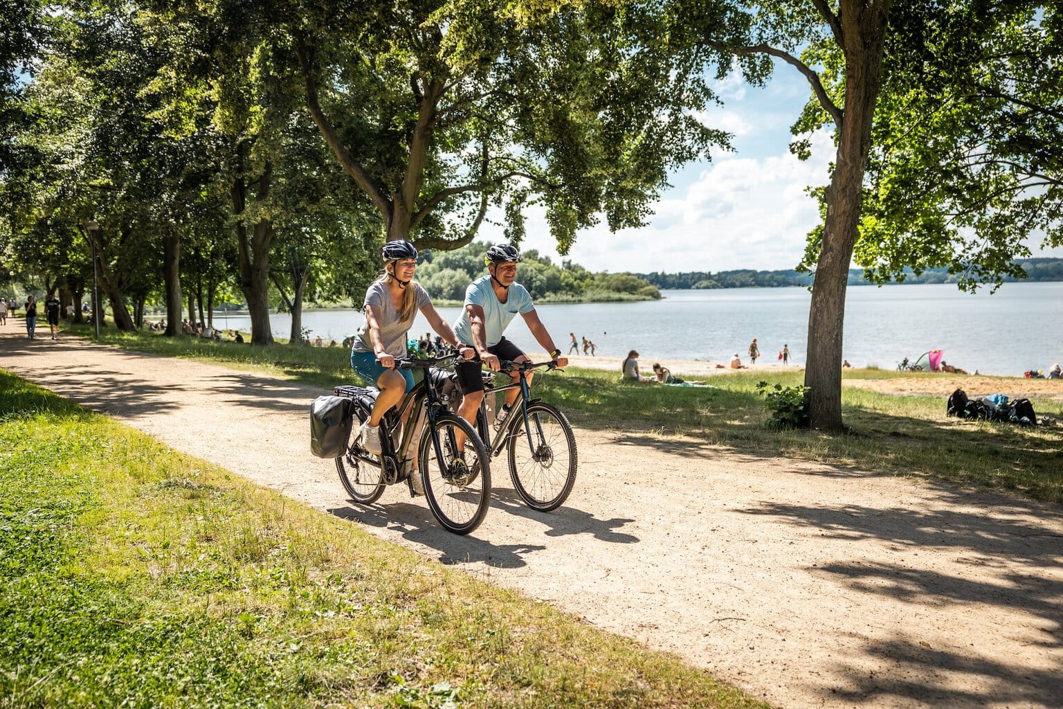 Twee fietsers rijden over het fietspad langs het strand van het stuwmeer in de fietsregio Bautzen. Op de achtergrond zie je het stuwmeer en mensen die op het strand van de zon genieten.