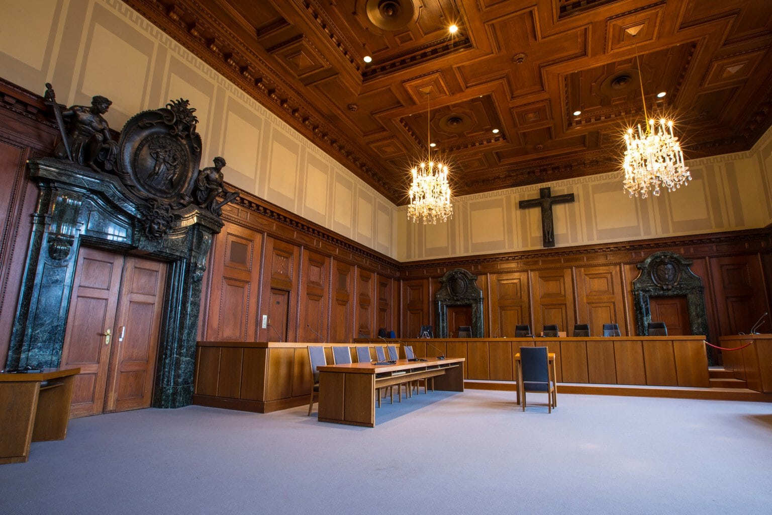 De rechtszaal 600 van de Neurenbergse processen in Neurenberg. Een grote ruimte met op de achtergrond een houten podium. Houten stoelen met grijze bekleding staan rond een houten tafel. Op de achtergrond staat een groot stenen kruis en aan het houten plafond hangen kroonluchters.