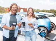 Twee mensen staan met een kopje koffie en een bagel in hun hand bij een wegrestaurant in Duitsland. Achter hen staat een elektrische auto op te laden.