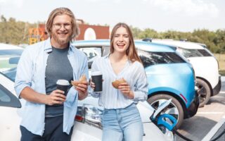 Twee mensen staan met een kopje koffie en een bagel in hun hand bij een wegrestaurant in Duitsland. Achter hen staat een elektrische auto op te laden.